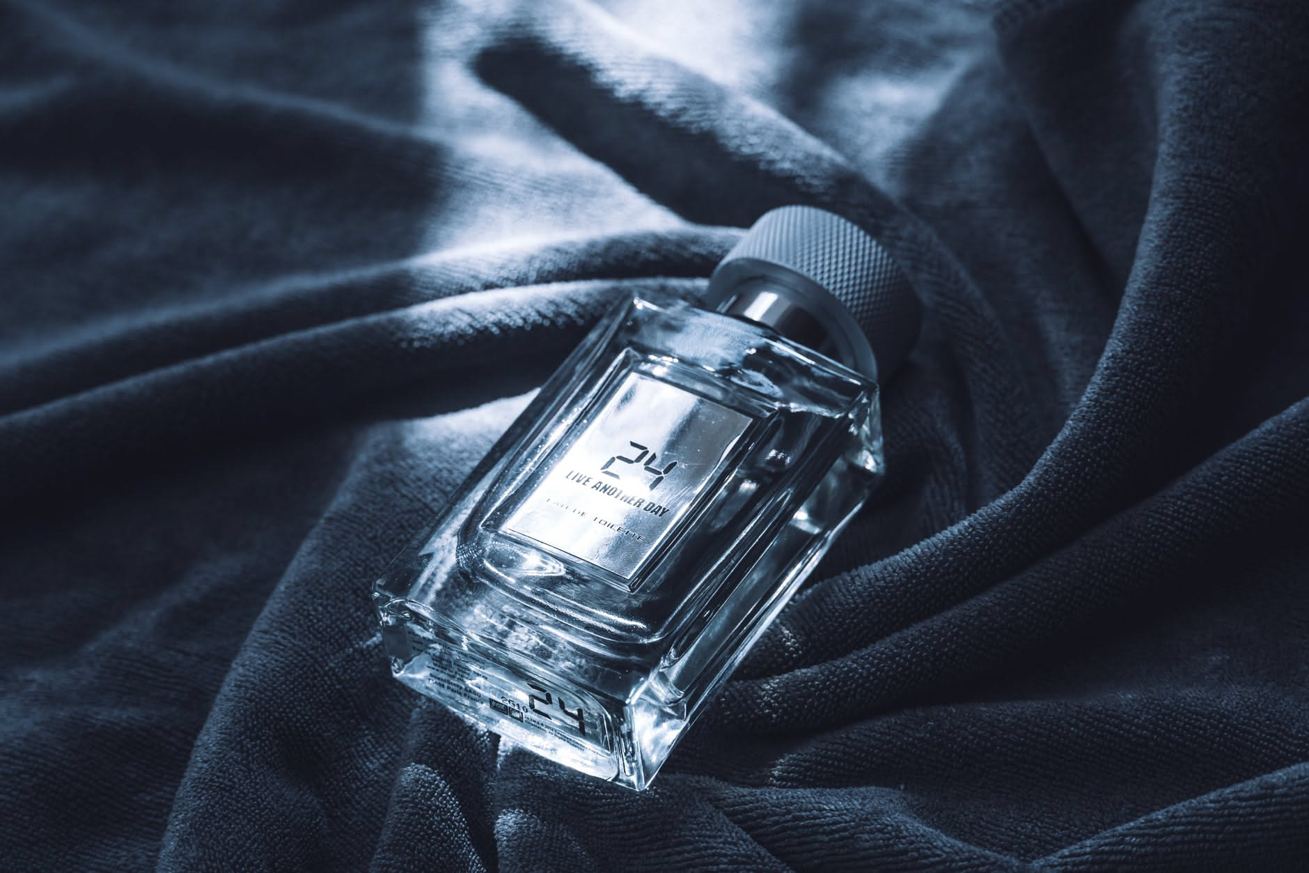 Flacone di profumo in vetro trasparente con liquido ambrato chiaro, riposto accanto a una finestra con luce naturale, con confezione originale sfocata sullo sfondo