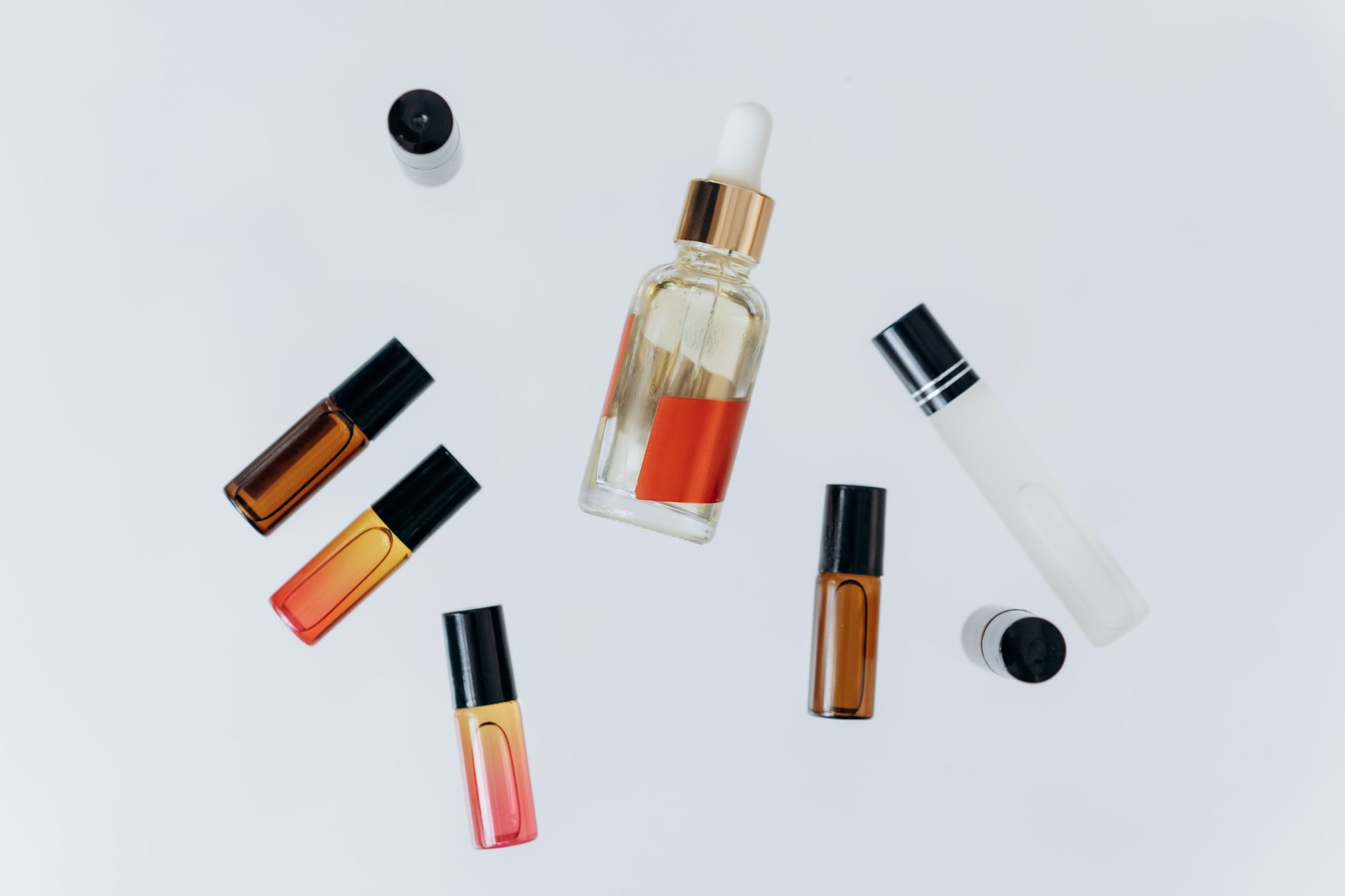Flaconi di profumo di diverse tonalità e colori disposti su una superficie bianca, con uno spray che nebulizza una fragranza trasparente