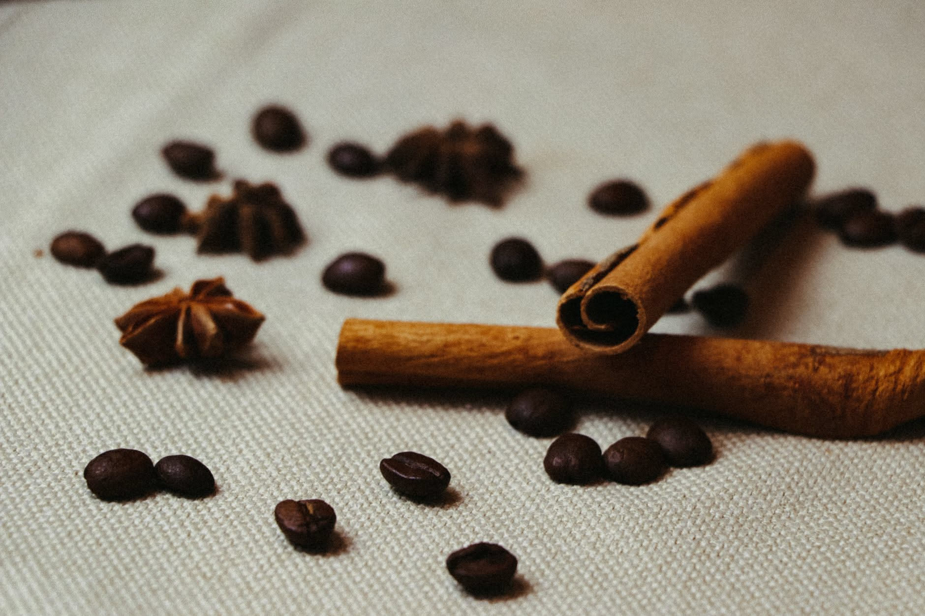 Bottiglia di profumo trasparente con liquido ambrato circondata da bastoncini di vaniglia, fave di cacao e spezie su sfondo neutro e luminoso